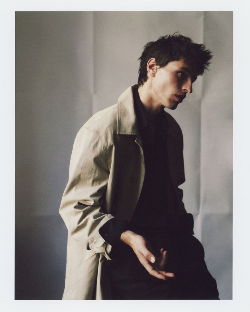 Portrait of a young adult male wearing a tan trench coat against a textured, neutral background in a photo studio setting.