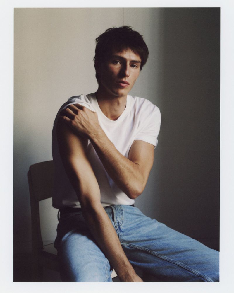 Clément Laguardia seated in a white t-shirt and light wash denim jeans against a muted neutral wall.