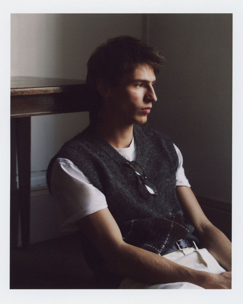 A young adult male sits indoors against a neutral wall, wearing a vest, white shirt, and light pants, with eyeglasses on his vest.