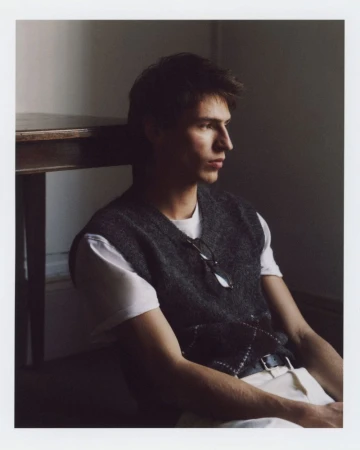 A young adult male sits indoors against a neutral wall, wearing a vest, white shirt, and light pants, with eyeglasses on his vest.