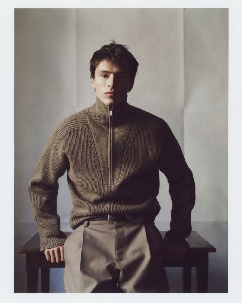 Indoor portrait of a young adult male wearing a zip-up sweater and trousers leaning against a wooden table with a neutral background.