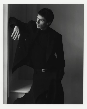 Model Clément Laguardia wearing a tailored black suit and turtleneck, leaning by a window in a black and white portrait.