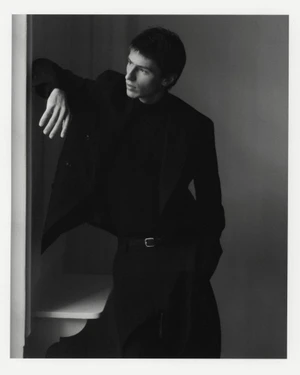 Model Clément Laguardia wearing a tailored black suit and turtleneck, leaning by a window in a black and white portrait.