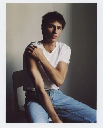 Clément Laguardia seated in a white t-shirt and light wash denim jeans against a muted neutral wall.