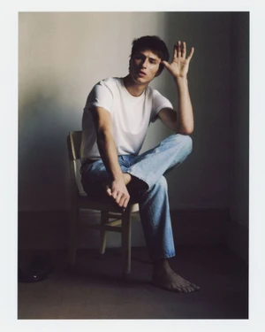 Young man in a white t-shirt and blue jeans sitting on a wooden chair against a plain wall for Zara Origins Fall 2025.