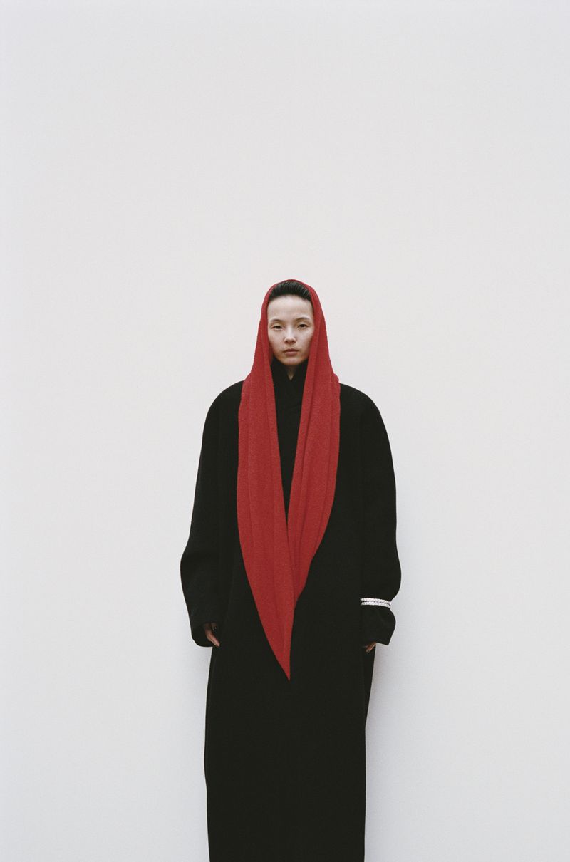 Portrait of a woman in a black coat and red headscarf against a plain white background, simple fashion with elegant style.