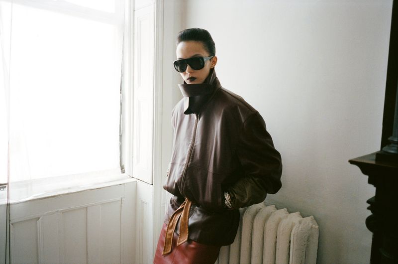An adult female poses indoors in a brown leather jacket, dark sunglasses, and a brown skirt, illuminated by natural light near a window.