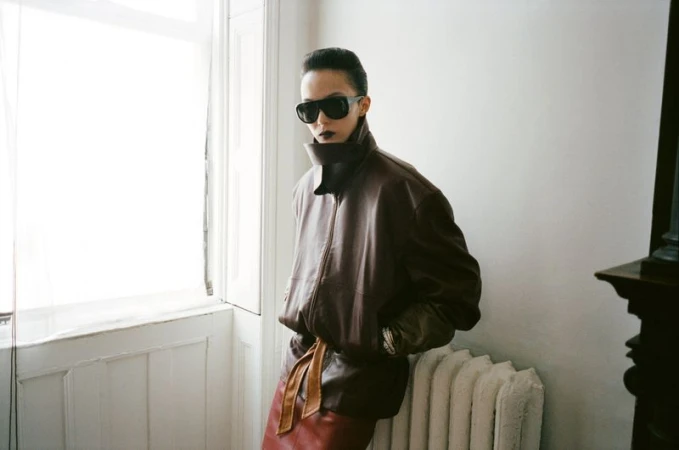 An adult female poses indoors in a brown leather jacket, dark sunglasses, and a brown skirt, illuminated by natural light near a window.