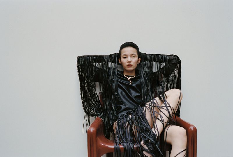 A stylish adult woman in a chair, wearing a fashionable black fringed outfit and gold necklace, poses against a plain background.