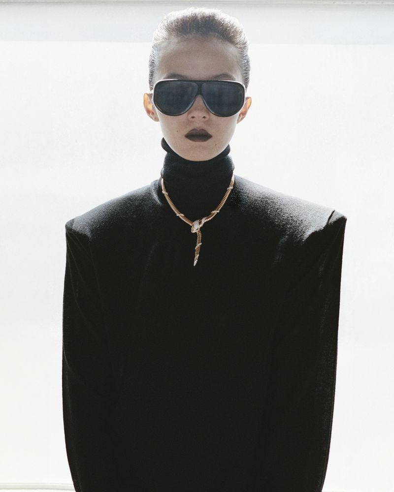 Fashionable adult female with sunglasses, black turtleneck sweater, and necklace posing indoors in front of white wall and window.