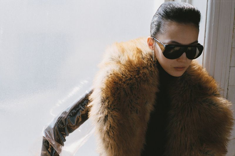 Stylish woman wearing sunglasses and a fur coat, posing indoors in front of a frosted glass window with natural lighting.