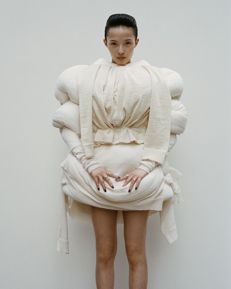 Asian fashion model wears oversized white padded clothing in a studio portrait against a neutral backdrop, looking directly at the camera.