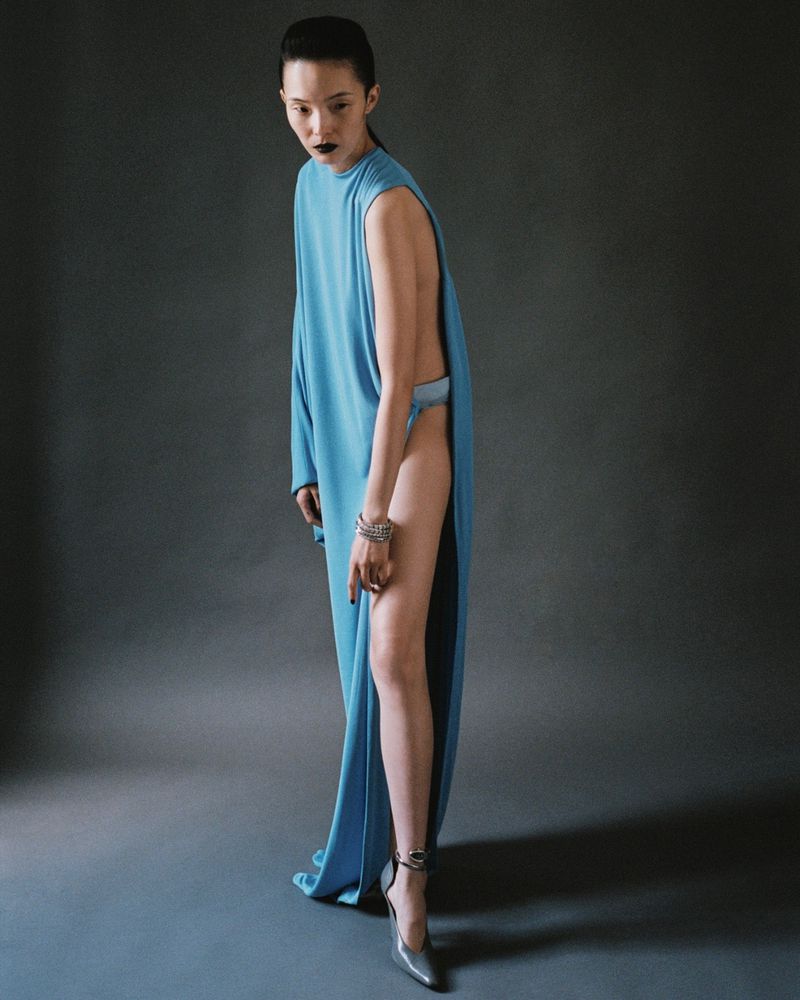 Woman in light blue gown with a side slit poses in front of a dark grey backdrop. Silver heels and jewelry complete the look.