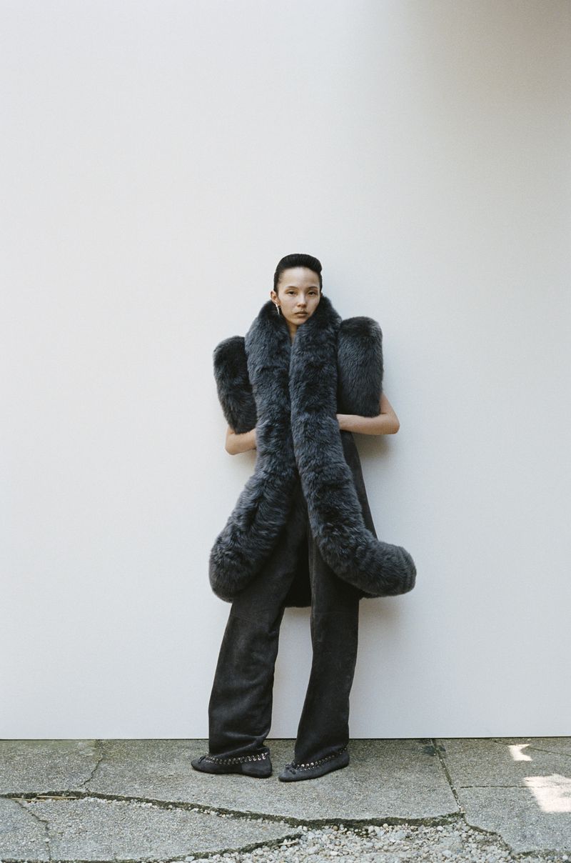 A slender Asian woman poses in flared gray pants and a large gray fur stole against a plain, bright wall. She wears dark studded flats.