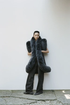 A slender Asian woman poses in flared gray pants and a large gray fur stole against a plain, bright wall. She wears dark studded flats.