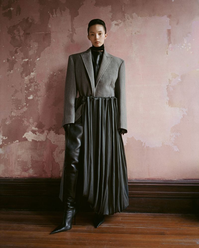 An Asian woman stands in a fashion-forward grey blazer and full grey skirt with black leather thigh-high boots against a textured wall.