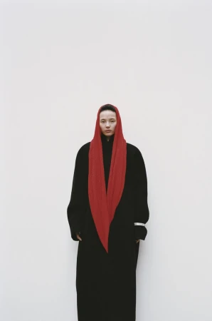 Xiao Wen Ju standing against a plain white backdrop wearing an ankle-length black coat and a vibrant red draped headscarf.