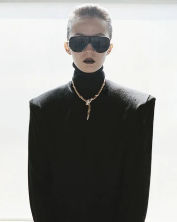 Model Xiao Wen Ju wearing black sunglasses, a gold snake necklace, and a black high-neck sweater with structured shoulders.