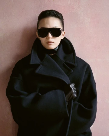 Model Xiao Wen Ju wearing oversized black tailored coat and black shield sunglasses against a textured dusty pink wall