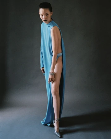 Model Xiao Wen Ju posing in a sleeveless sky blue open-sided draped gown and silver heels against a grey studio backdrop.