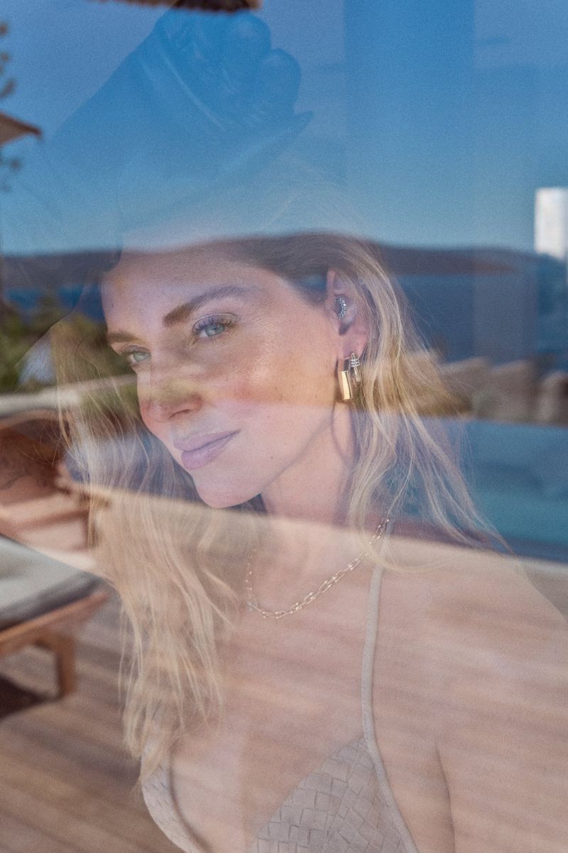 Portrait through a window shows a woman with blonde hair wearing gold jewelry and a light beige top, reflected against a landscape backdrop.
