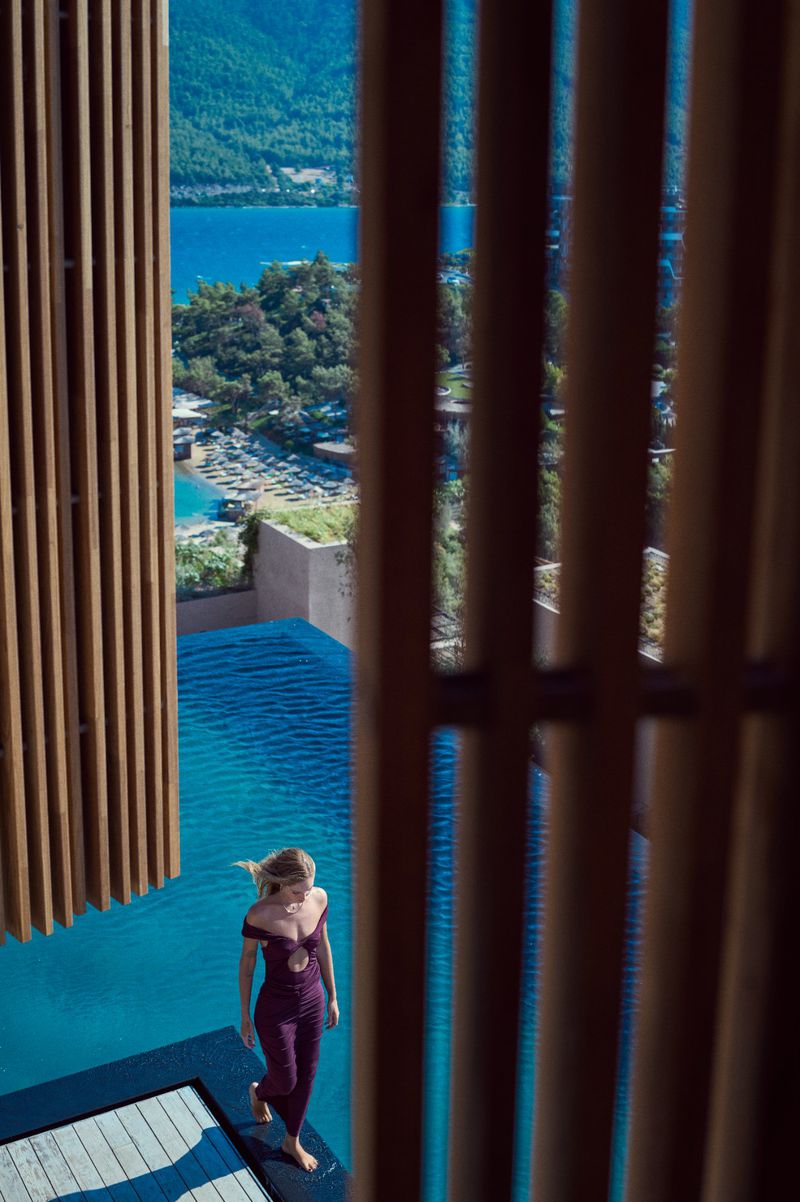 Woman in red jumpsuit standing near turquoise pool with ocean and green mountains in background, seen through wood slats. Luxury travel.