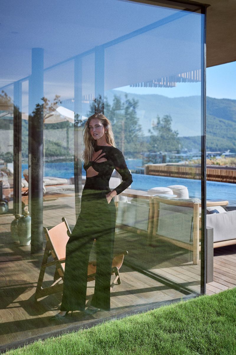 Woman in a cutout top and wide-leg pants poses behind a large glass window, reflecting a luxury pool and hillside view.
