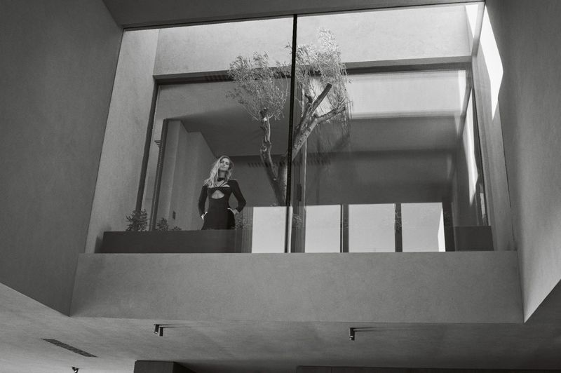 Elegant blonde woman in a modern black dress posing near a large window with a tree inside a contemporary architectural space.