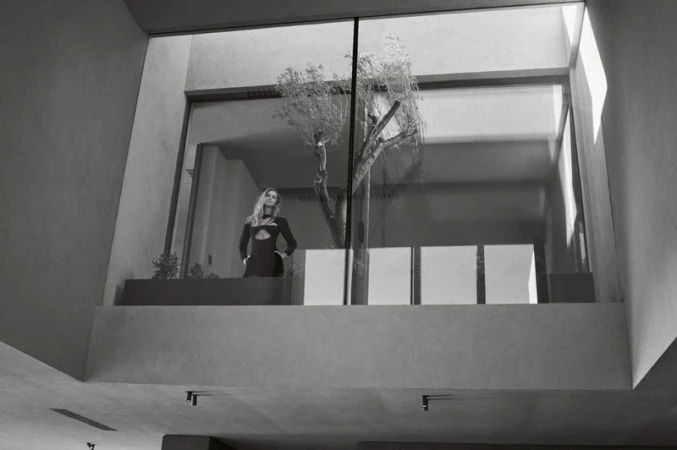 Elegant blonde woman in a modern black dress posing near a large window with a tree inside a contemporary architectural space.