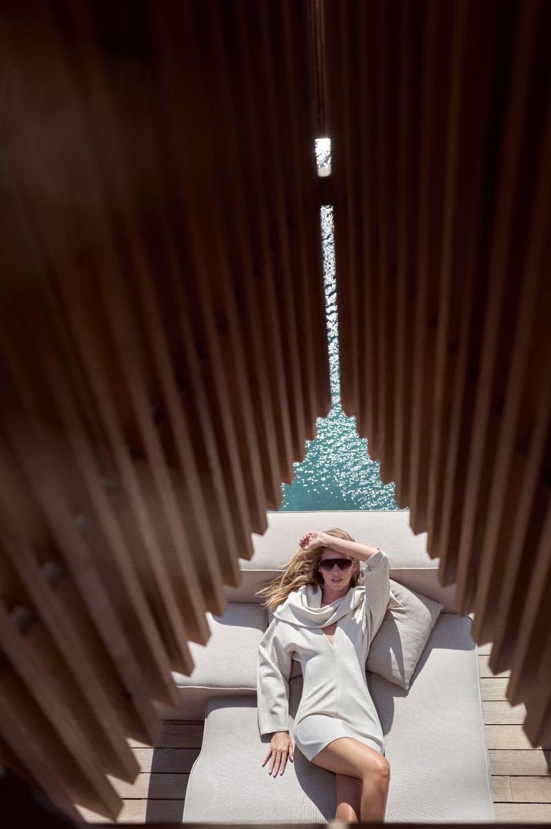 Woman in a cover-up lounging on an outdoor sofa under a wooden structure, with a view of blue water in the background. Sunglasses protect her eyes.