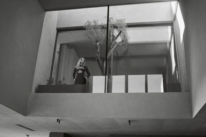 Chiara Ferragni in a black cutout dress posing on an upper interior glass balcony beside an indoor courtyard tree in black and white.