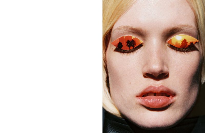 Close-up of a blonde woman's face with landscape-themed eye makeup featuring palm trees and buildings in silhouette against an orange sunset.