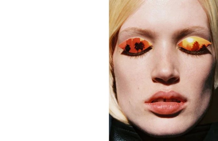 Close-up of a blonde woman's face with landscape-themed eye makeup featuring palm trees and buildings in silhouette against an orange sunset.