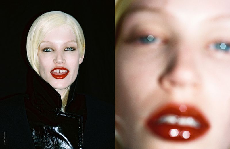 Close-up of a fair-skinned woman with platinum blonde hair, red lipstick, and black jacket on a dark background.