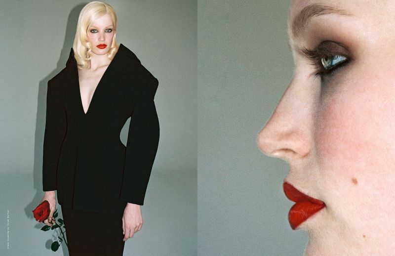 Woman with pale blonde hair in a black jacket holds a red rose in a studio setting, side face with red lipstick in frame.