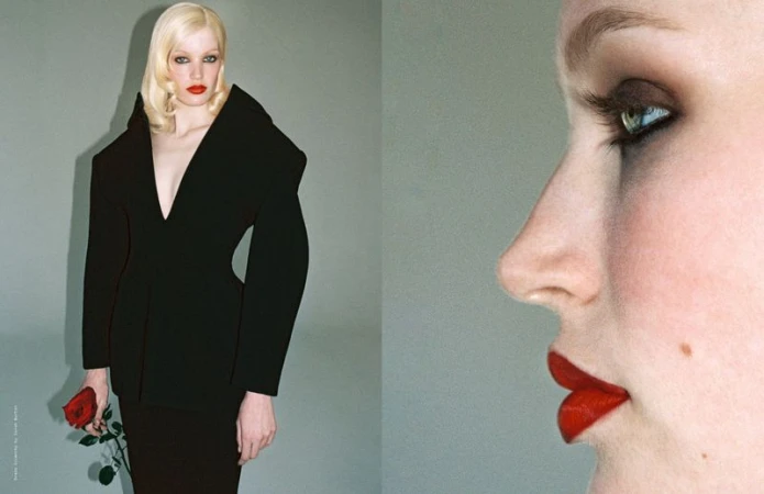 Woman with pale blonde hair in a black jacket holds a red rose in a studio setting, side face with red lipstick in frame.