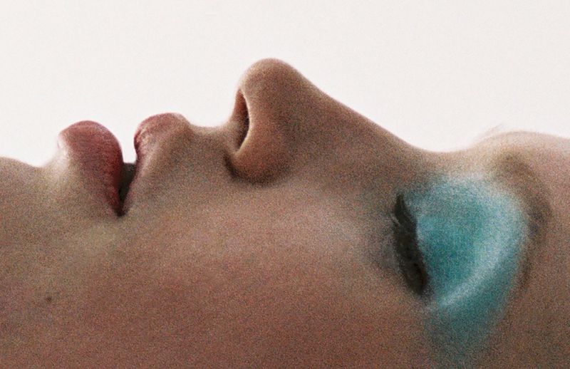 Close-up of a person's face, focusing on the nose and mouth with a hint of blue makeup under the eye, set against a light background.