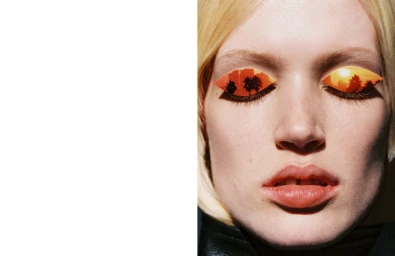 Close-up beauty portrait of a blonde woman with paper sunset collage cutouts over her closed eyes, set against white negative space.