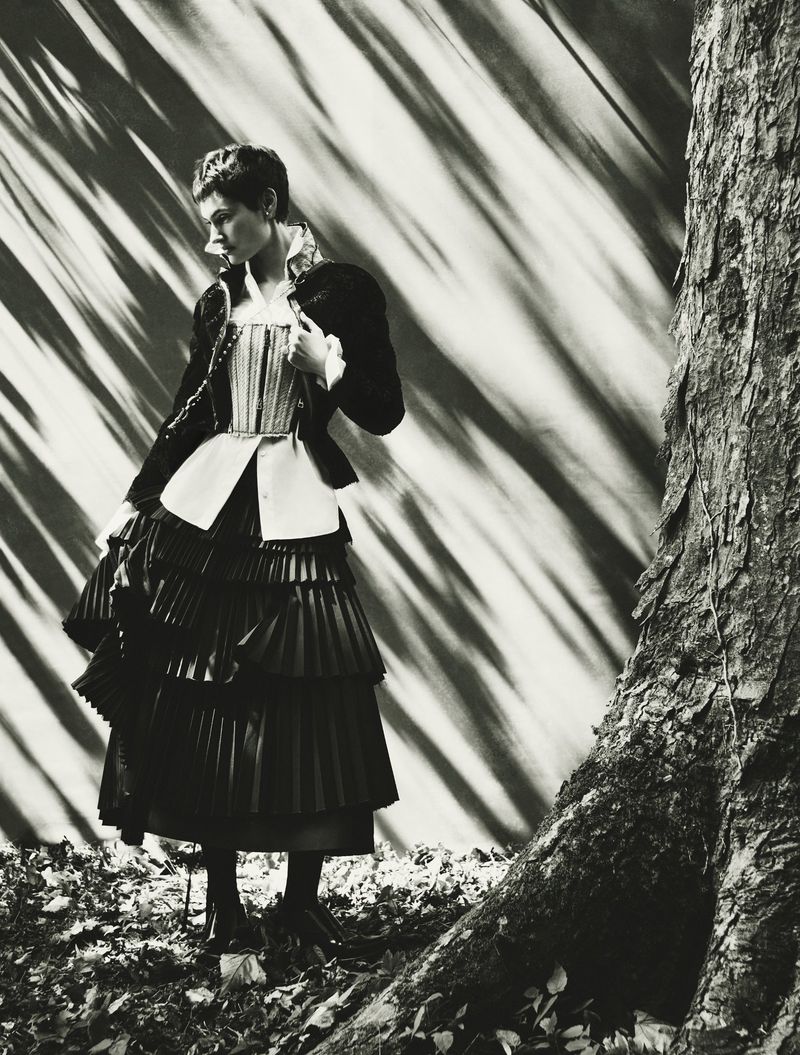 Monochrome shot of a woman in a tiered dress holding a hat standing in front of a patterned backdrop next to a textured tree.