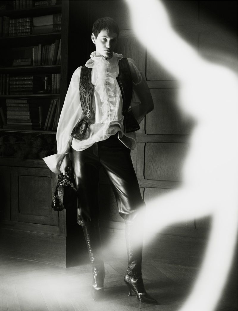 Monochrome fashion shot of a person wearing a ruffled shirt, vest, leather pants, and heeled boots posing indoors with shelves in the background.