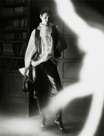 Monochrome fashion shot of a person wearing a ruffled shirt, vest, leather pants, and heeled boots posing indoors with shelves in the background.