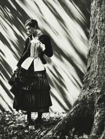 Model Greta Hofer wearing a corseted ruffled jacket and tiered pleated skirt next to a tree trunk in monochrome lighting.