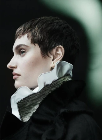 Close-up profile portrait of a woman with short dark hair wearing a dark coat and high white ruffled collar against a dark background.