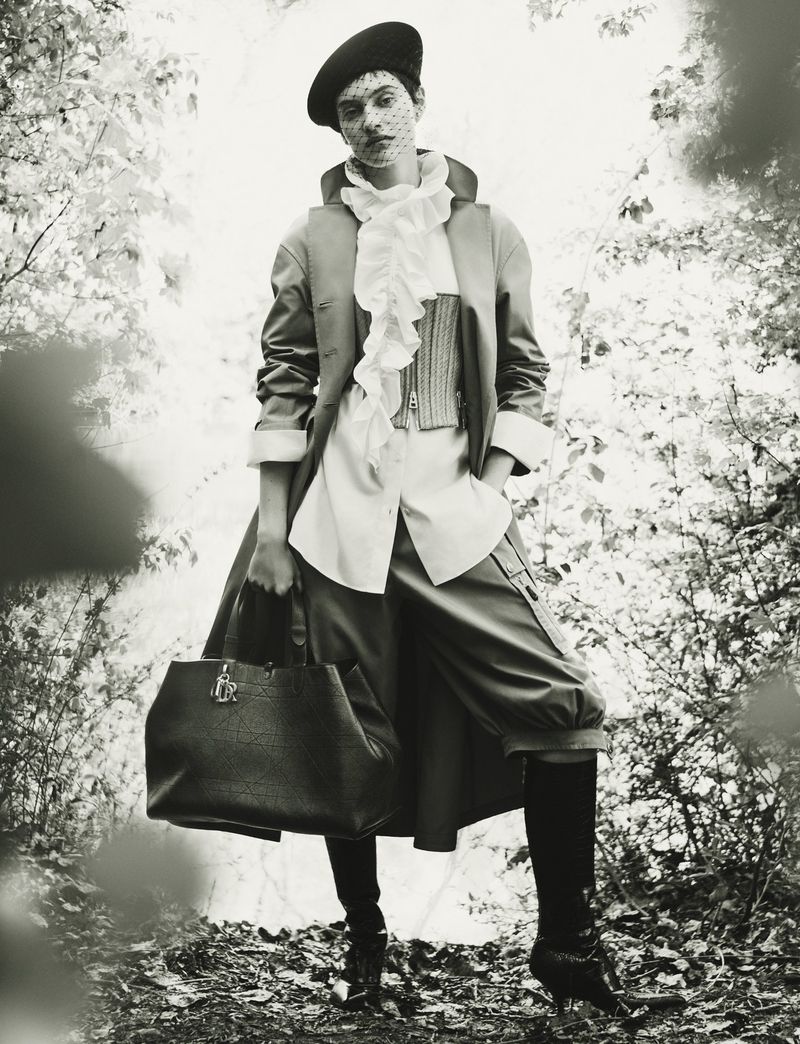 Model in beret with veil, ruffled collar shirt, coat, and carrying a large dark handbag standing outdoors among trees.