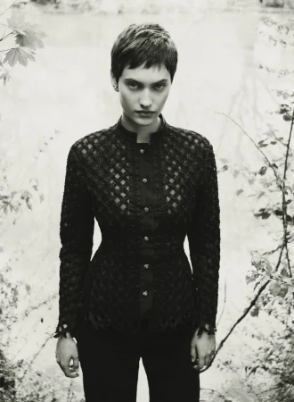 Model with short dark hair wearing a patterned black buttoned top outdoors against a blurred foliage background.
