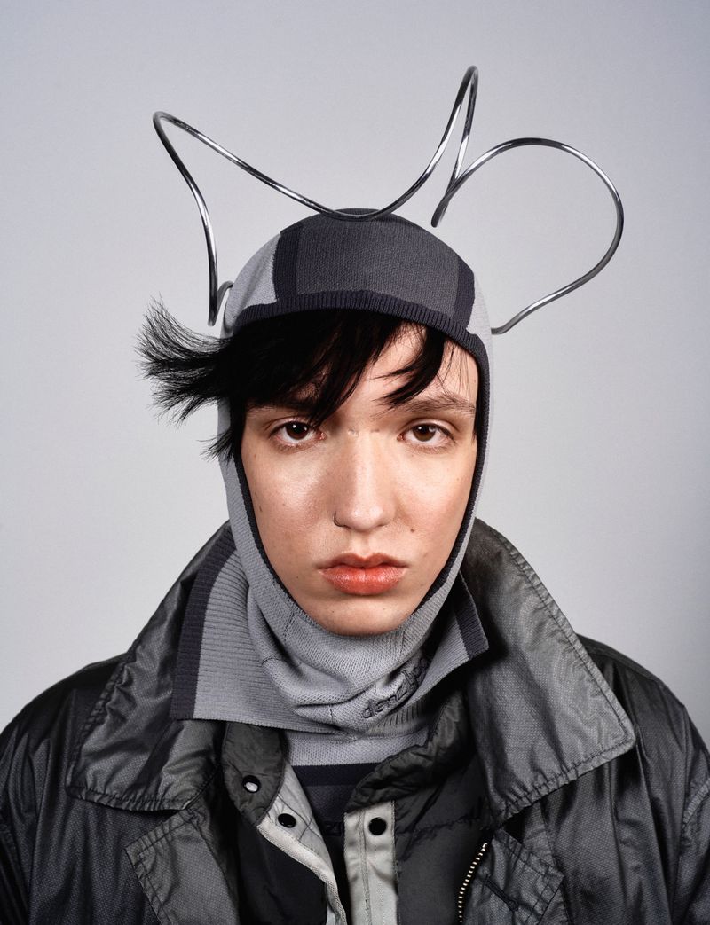 Portrait of a person wearing a gray knit balaclava and a dark gray shiny jacket against a light gray background in a studio.