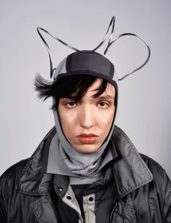 Portrait of a person wearing a gray knit balaclava and a dark gray shiny jacket against a light gray background in a studio.
