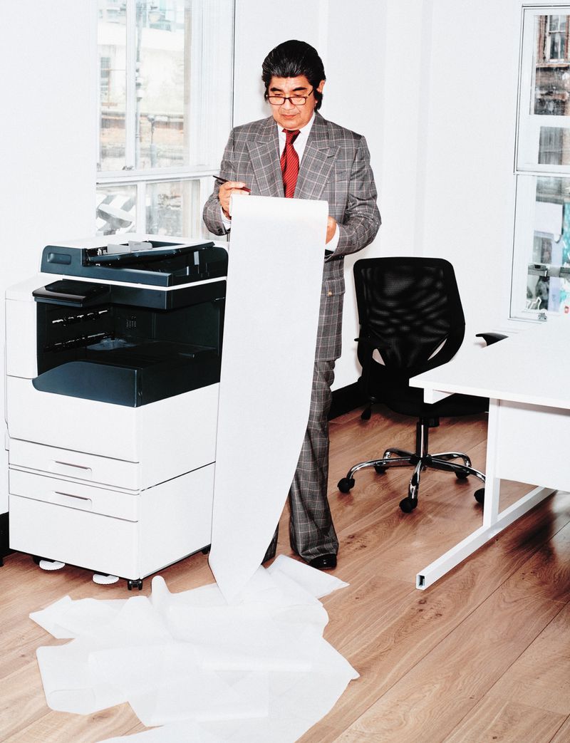 Mature businessman with eyeglasses in a gray suit stands by a copy machine holding a long printout in an office with a wooden floor.