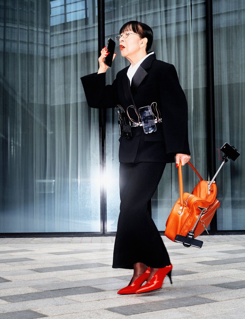 Adult woman in a black suit and red heels using a smartphone outdoors with office buildings in the background on a sunny day.