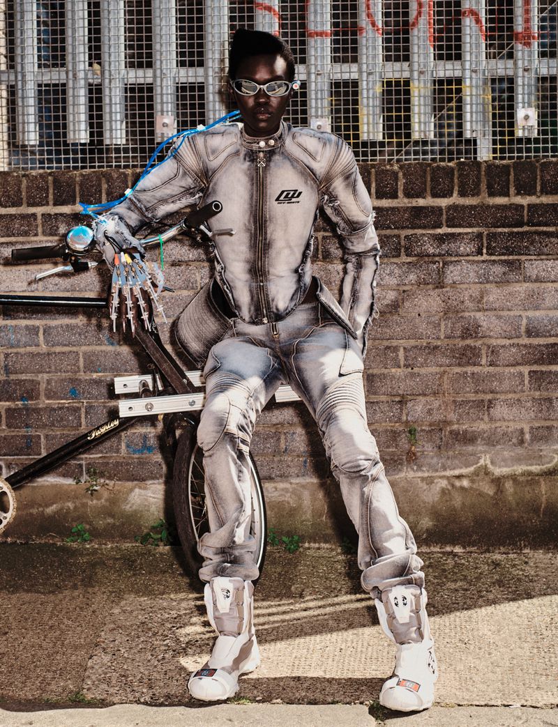 A Black person wearing a futuristic suit and glasses, leaning on a bicycle with metallic accents against a brick wall and metal fence.